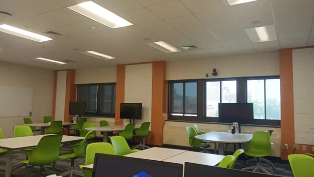 An active learning classroom with round tables, green chairs, and mobile monitor stands positioned near the windows for group work.