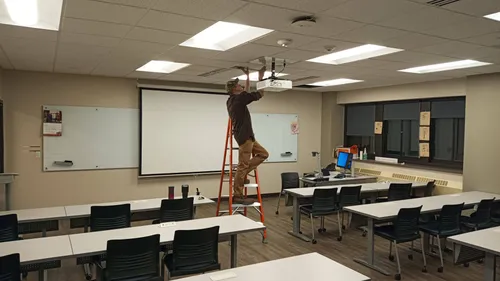A technician on a ladder installs a ceiling-mounted projector above a classroom with a pull-down screen and rows of tables.