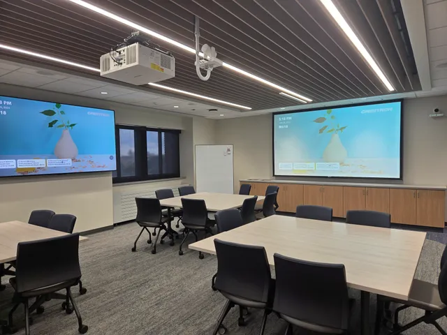 A conference facility with two 100-inch flatscreens showing a wireless presentation interface, a wall-mounted conference camera, and a ceiling-mounted projector.