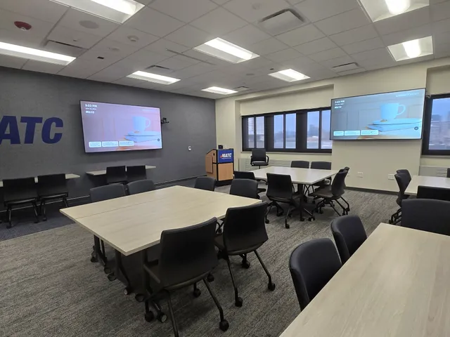 A conference room with movable tables and chairs, a lectern, and two large wall-mounted displays showing a wireless presentation screen.