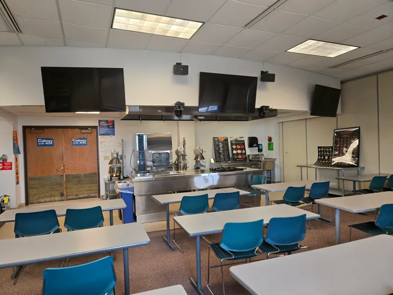 A culinary arts classroom with a stainless steel demonstration kitchen and three flat-panel displays mounted above the cooking equipment.