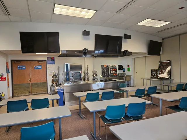 A culinary arts classroom with a stainless steel demonstration kitchen and three flat-panel displays mounted above the cooking equipment.