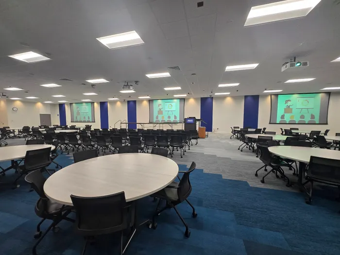 A large event center with round tables, rolling chairs, and three wall-mounted projection screens displaying a presentation slide.