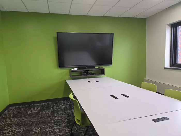 A small huddle room with a green accent wall, a large wall-mounted display, and a media shelf feeding a conference table.