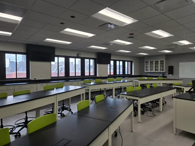 A science lab classroom with lab benches, green task chairs, and two large flat-panel displays mounted in the ceiling corners.