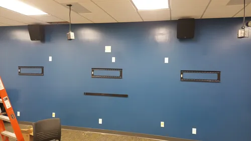 Three empty wall mounting brackets and ceiling speakers installed on a blue accent wall, with a ladder still in the room ahead of the displays being hung.